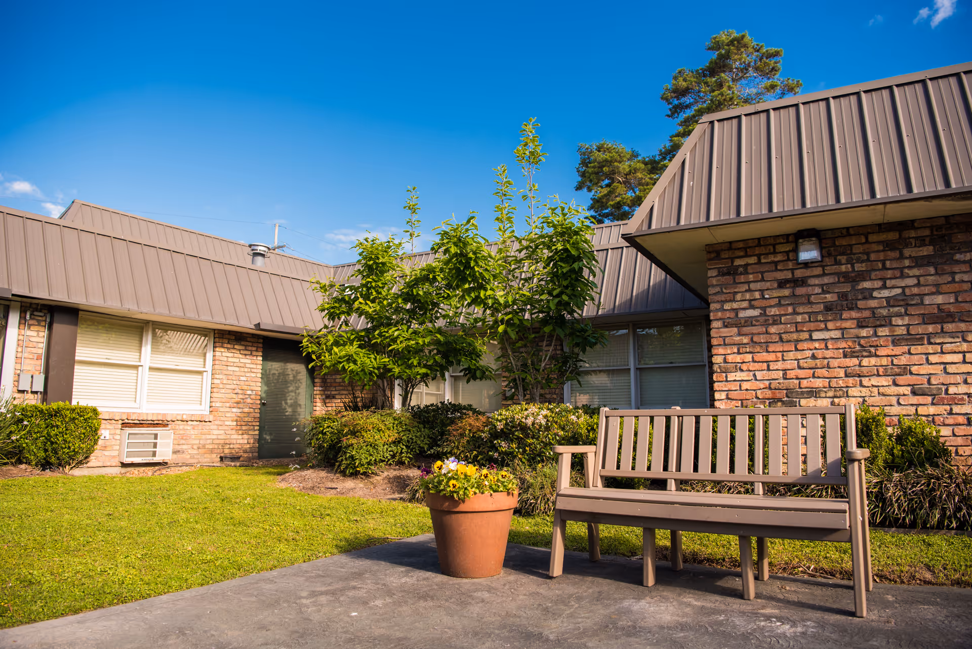 Outdoor view of a single-story brick building with a bench, potted flowers, shrubs, and a clear blue sky.