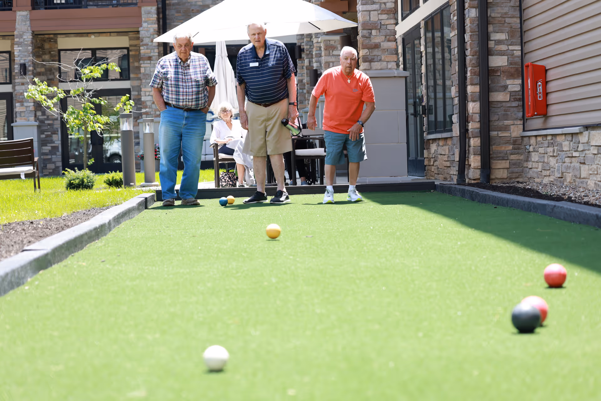 Three elderly men playing bocce ball on a green outdoor court at a senior living facility, with a woman sitting under a white umbrella in the background near a stone building.