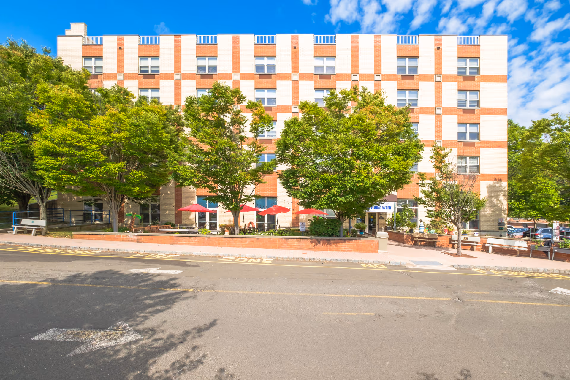 Multi-story brick-and-white senior living building front with trees, red patio umbrellas, and a street in the foreground.