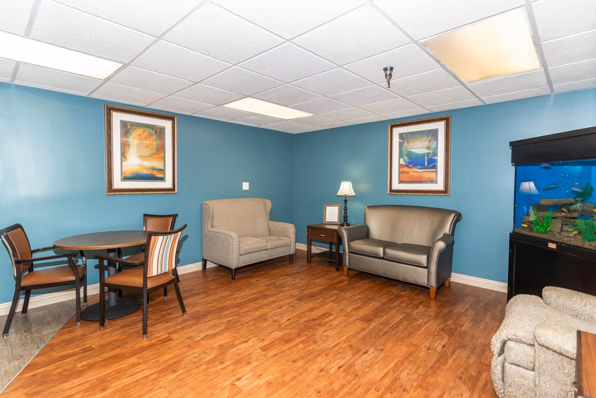 Small communal sitting room with a round table and chairs, two sofas, framed artwork, and a large fish tank against teal walls.