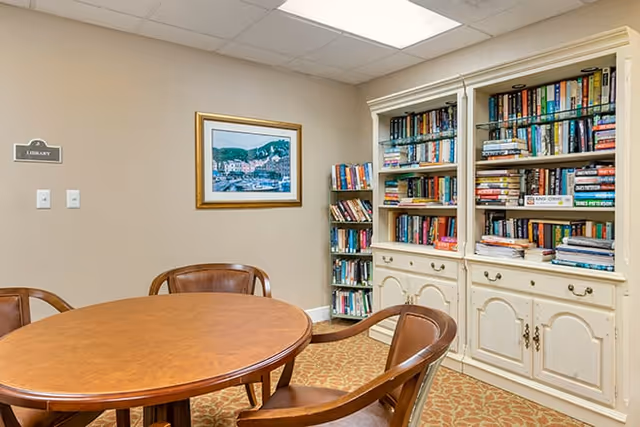 A small library room with a round wooden table surrounded by four wooden chairs. There are two white bookshelves filled with books against the wall, and a smaller bookshelf in the corner. A framed painting hangs on the beige wall above the bookshelves. A sign on the wall reads 'Library'.