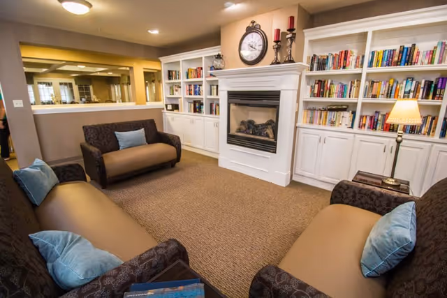 A cozy living room area with three brown sofas each adorned with light blue pillows, a white fireplace with a clock and candle holders on the mantel, and built-in white bookshelves filled with books. A table lamp is on a side table next to one of the sofas, and a large mirror is mounted on the wall behind one sofa.