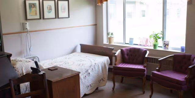 A bright senior bedroom with a single bed covered by a quilt, a wooden nightstand and dresser, two upholstered chairs by a large window, and framed photos on the wall.