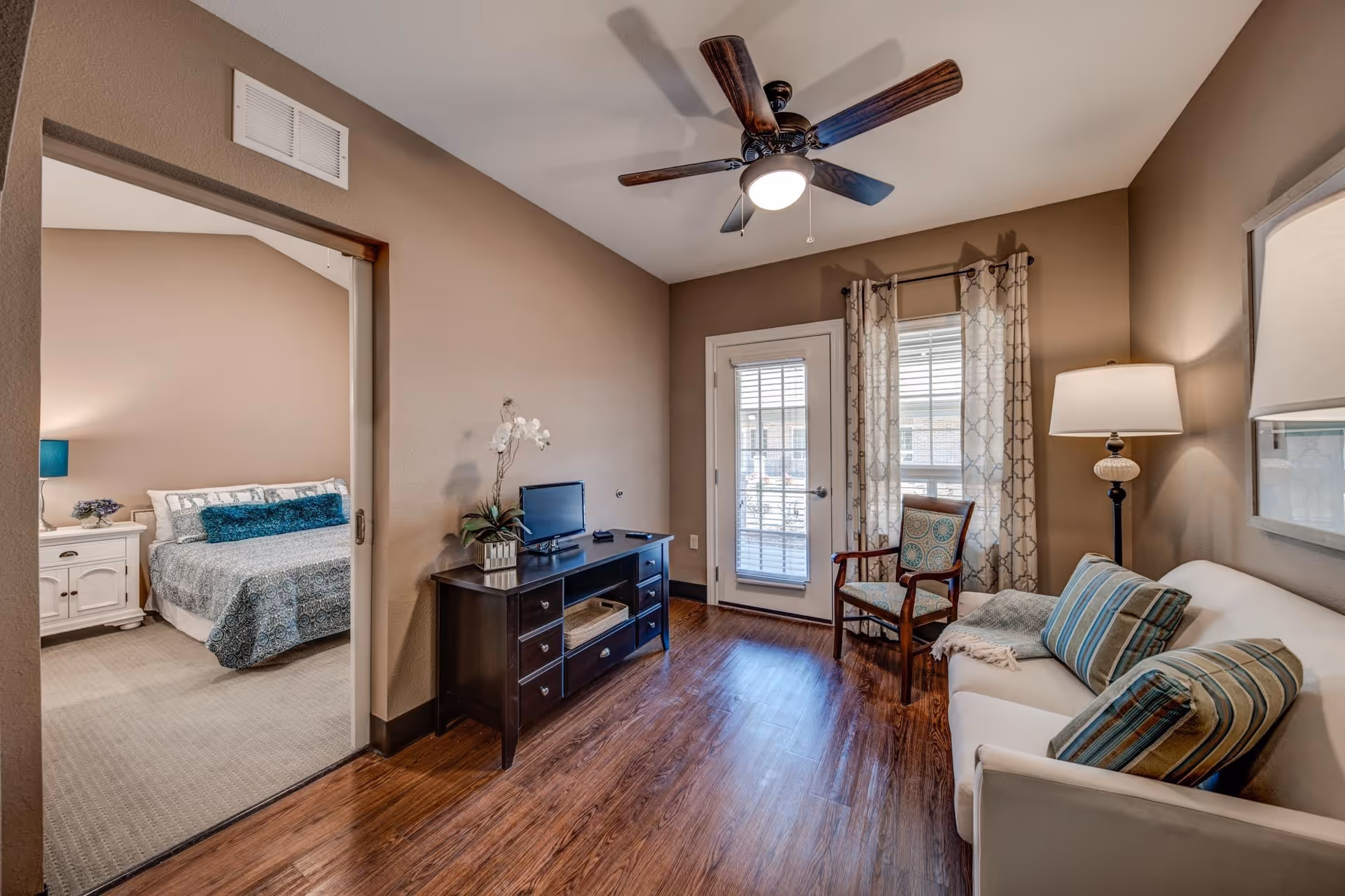 A cozy living room with wooden flooring, a ceiling fan, a white sofa with striped cushions, a floor lamp, a wooden chair with patterned upholstery, and a TV on a dark wooden stand. The room has a glass door with curtains leading outside. Adjacent to the living room is a bedroom with a bed, nightstand, and lamp visible through an open doorway.