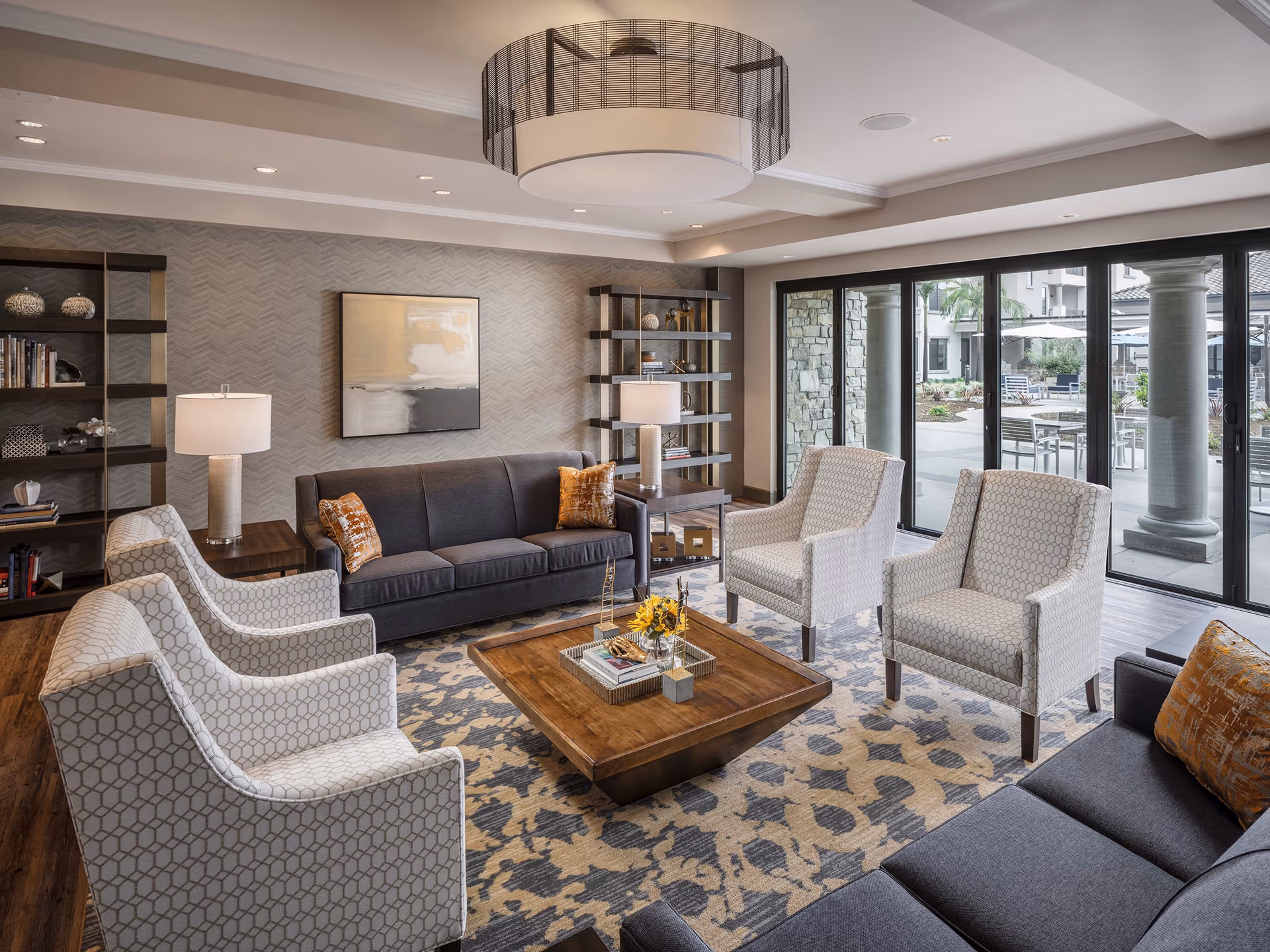 Bright, modern lounge with sofas, patterned armchairs, a wooden coffee table, shelving, and large sliding glass doors to an outdoor patio.