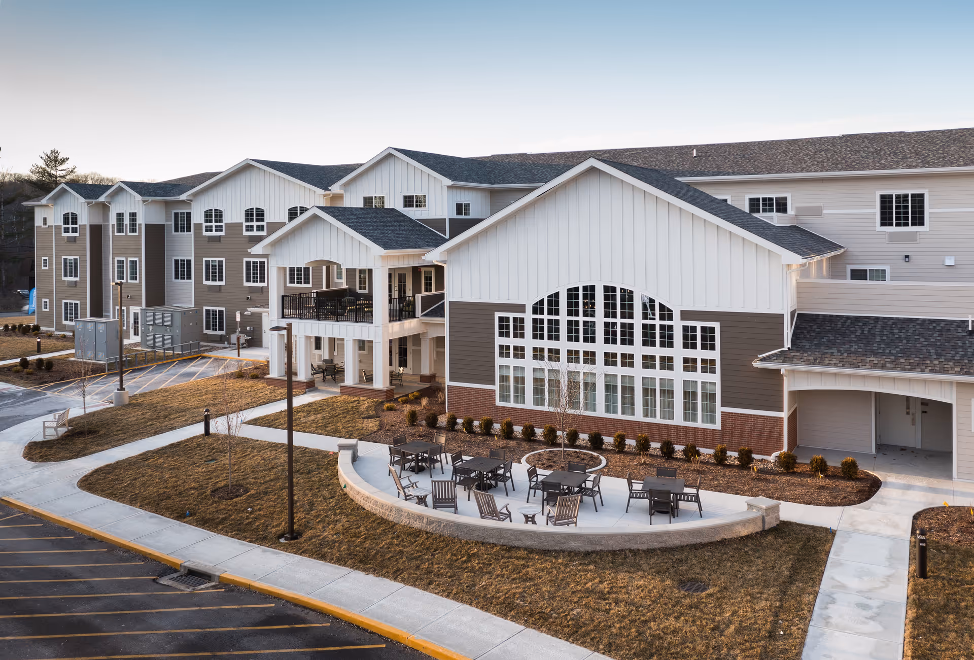 Exterior view of a modern senior living facility building with multiple windows, a covered balcony, and an outdoor seating area with tables and chairs arranged on a curved concrete patio. The surrounding area includes landscaped grass and paved walkways.