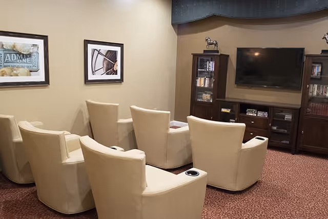 A cozy media room with six beige recliner chairs arranged in two rows facing a large flat-screen TV mounted on the wall. The room has a patterned red carpet, beige walls with two framed pictures, and dark wood cabinets holding books and decorative items.