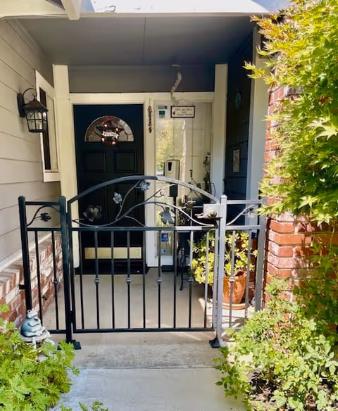 Front porch entrance of a senior living facility with a black decorative metal gate, a black door with a wreath, potted plants, and a brick wall partially covered with green foliage.