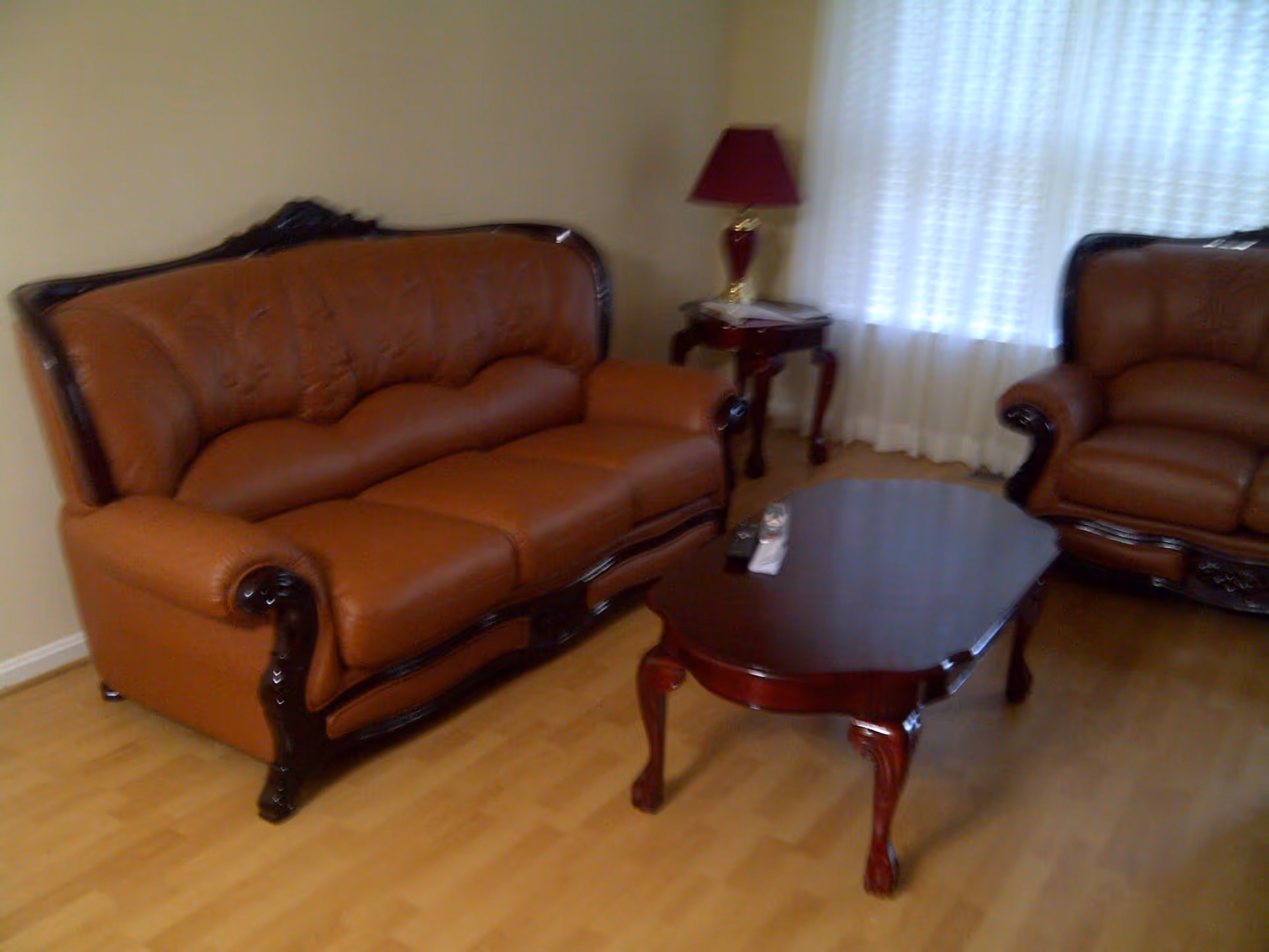 A living room with a brown leather sofa and matching armchair, a wooden coffee table with remote controls on it, a side table with a red lamp, and a window with white curtains.