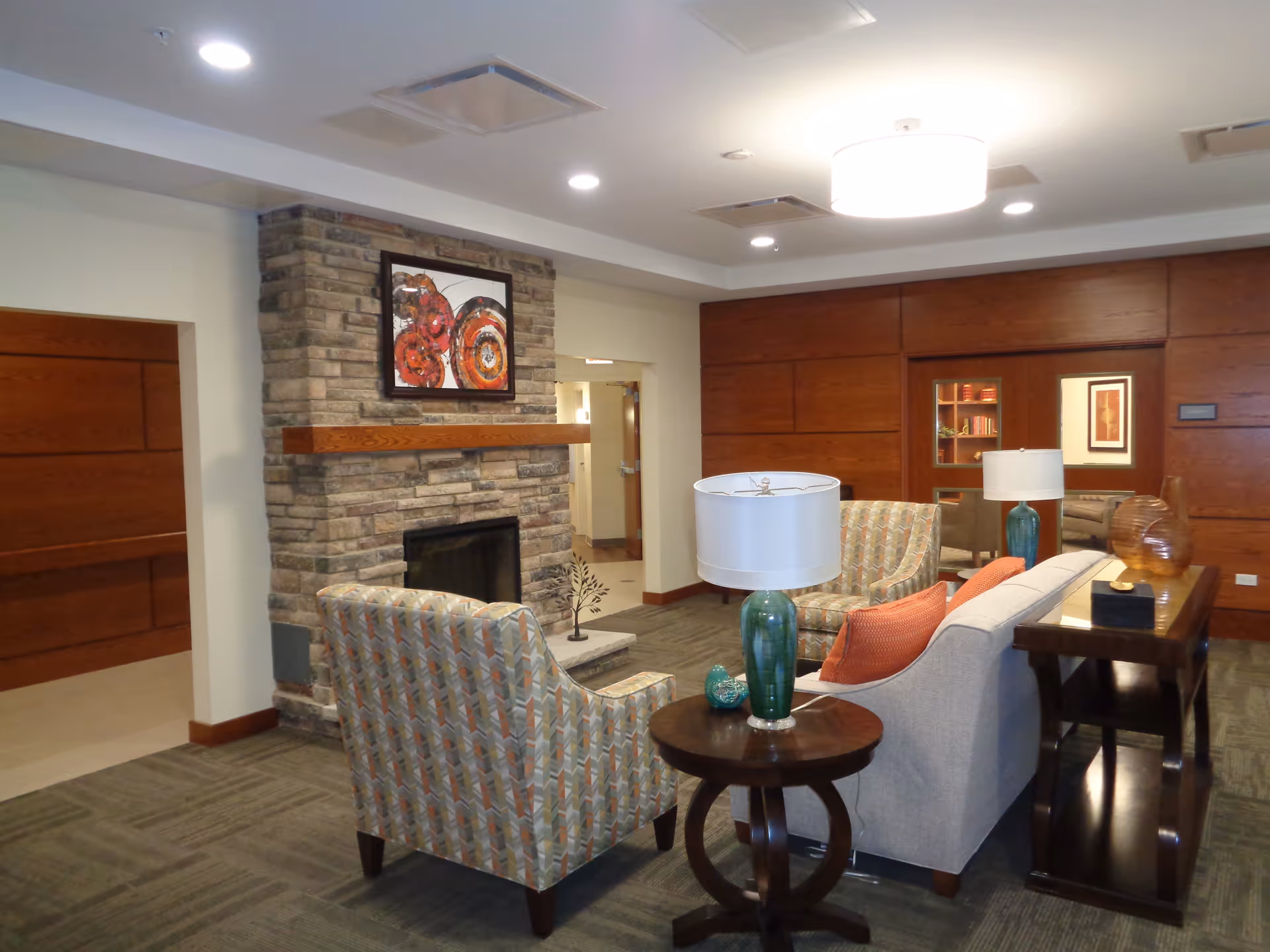 A cozy living room area with a stone fireplace, two patterned armchairs, a beige sofa with orange pillows, a round wooden side table with a green lamp, and a wooden console table with decorative items. The walls are wood-paneled and there is a framed abstract painting above the fireplace.