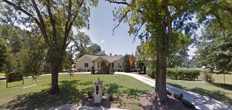 Single-story light-colored residential building with a covered front porch, lawn, mature trees, and a curved driveway leading to the entrance.