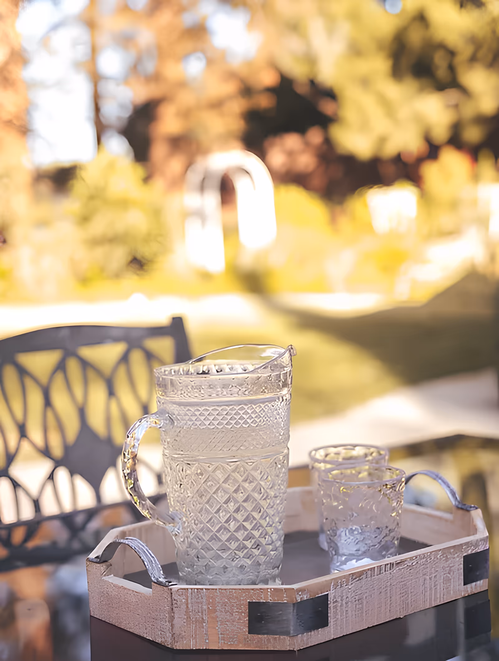 A clear glass pitcher filled with water and two matching clear glass tumblers on a wooden tray outdoors, with a metal chair and blurred garden background.