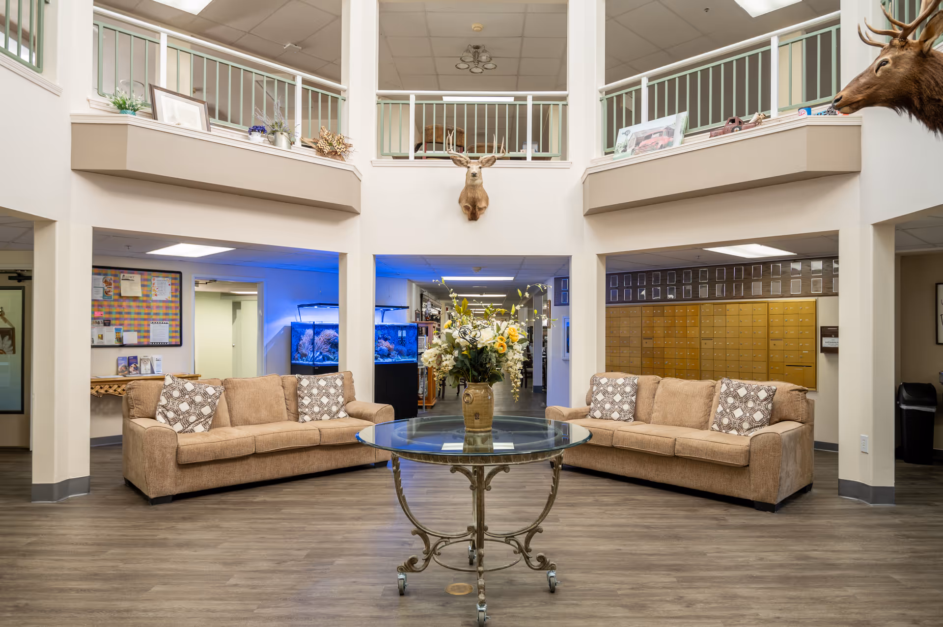 Interior view of a senior living facility lobby with two beige sofas facing each other, a round glass table with a floral arrangement in the center, mounted deer heads on the walls, and a set of mailboxes on the right side. There are aquariums with blue lighting in the background and a second-floor balcony with railings above.