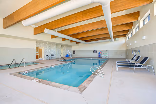 Indoor swimming pool area with clear blue water, surrounded by a beige tiled deck. There are several lounge chairs lined up against the right wall, and wooden beams with white pipes run across the ceiling. The far wall has a mosaic tile section with two chairs and a door, and there is a pool lift for accessibility.