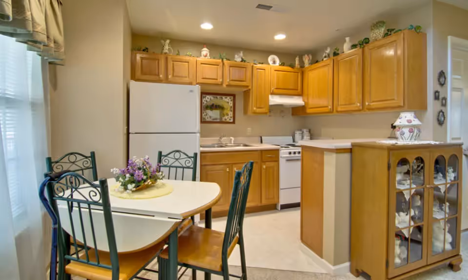 A cozy kitchen and dining area featuring wooden cabinets, a white refrigerator, a stove, and a small dining table with four chairs. The table has a floral centerpiece, and there is a glass-front cabinet displaying decorative items. The room is softly lit with recessed lighting and natural light from a window with curtains.
