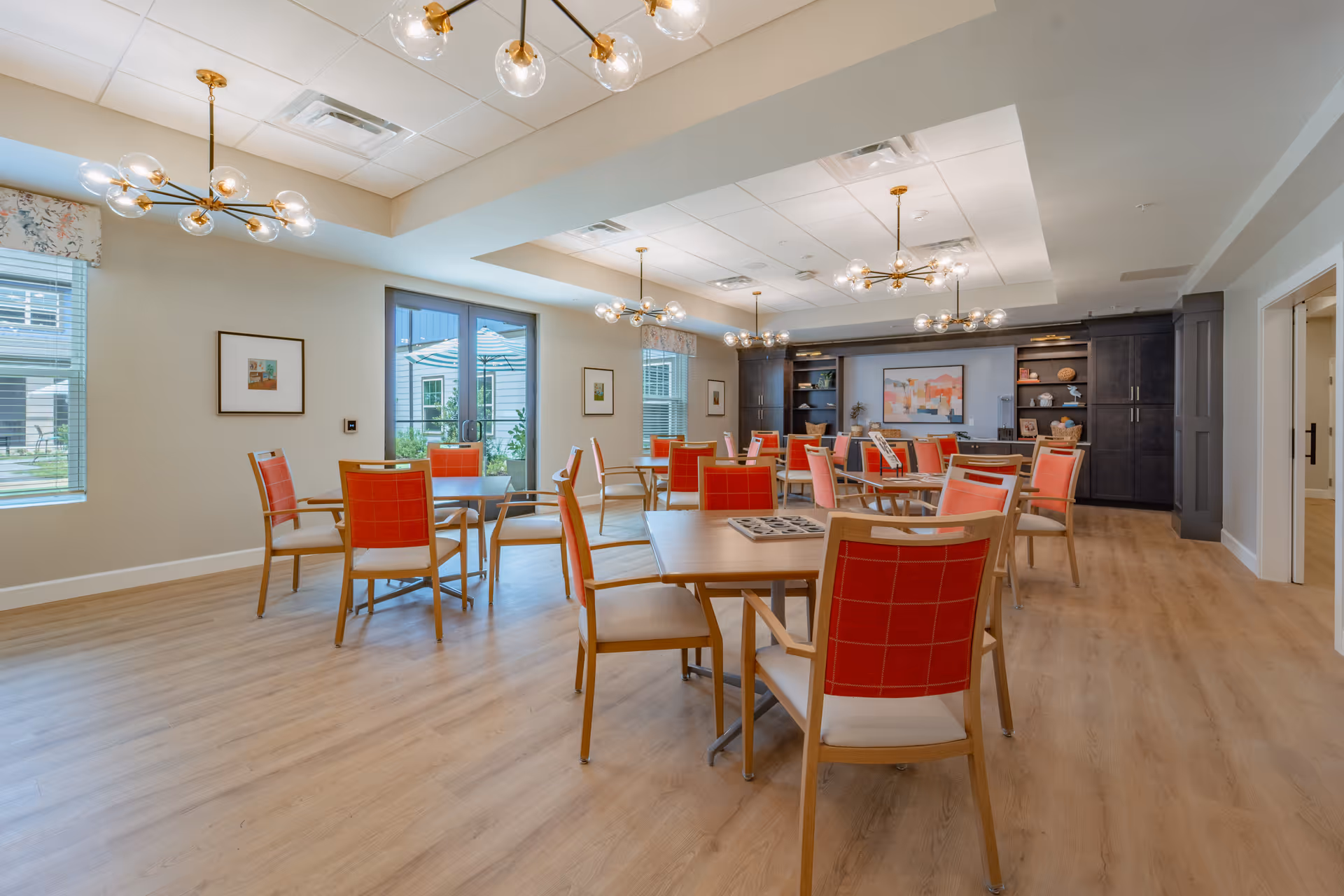 A bright and spacious dining room in a senior living facility with multiple wooden tables and chairs featuring red cushions. The room has large windows and glass doors letting in natural light, modern light fixtures hanging from the ceiling, and built-in dark wood cabinets along the back wall with decorative items and artwork.