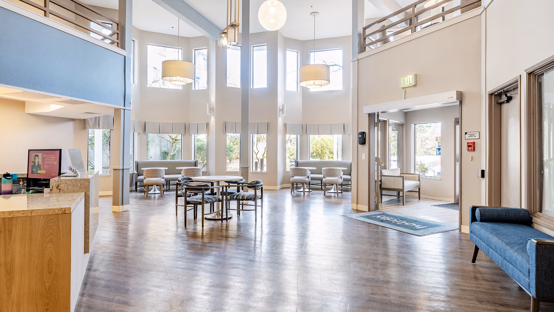 Bright and spacious senior living facility common area with high ceilings and large windows letting in natural light. The room features a round table with chairs in the center, seating areas with cushioned chairs and sofas along the walls, modern pendant lights hanging from the ceiling, and a reception desk on the left side. There is an exit door on the right side with a blue welcome mat in front of it.