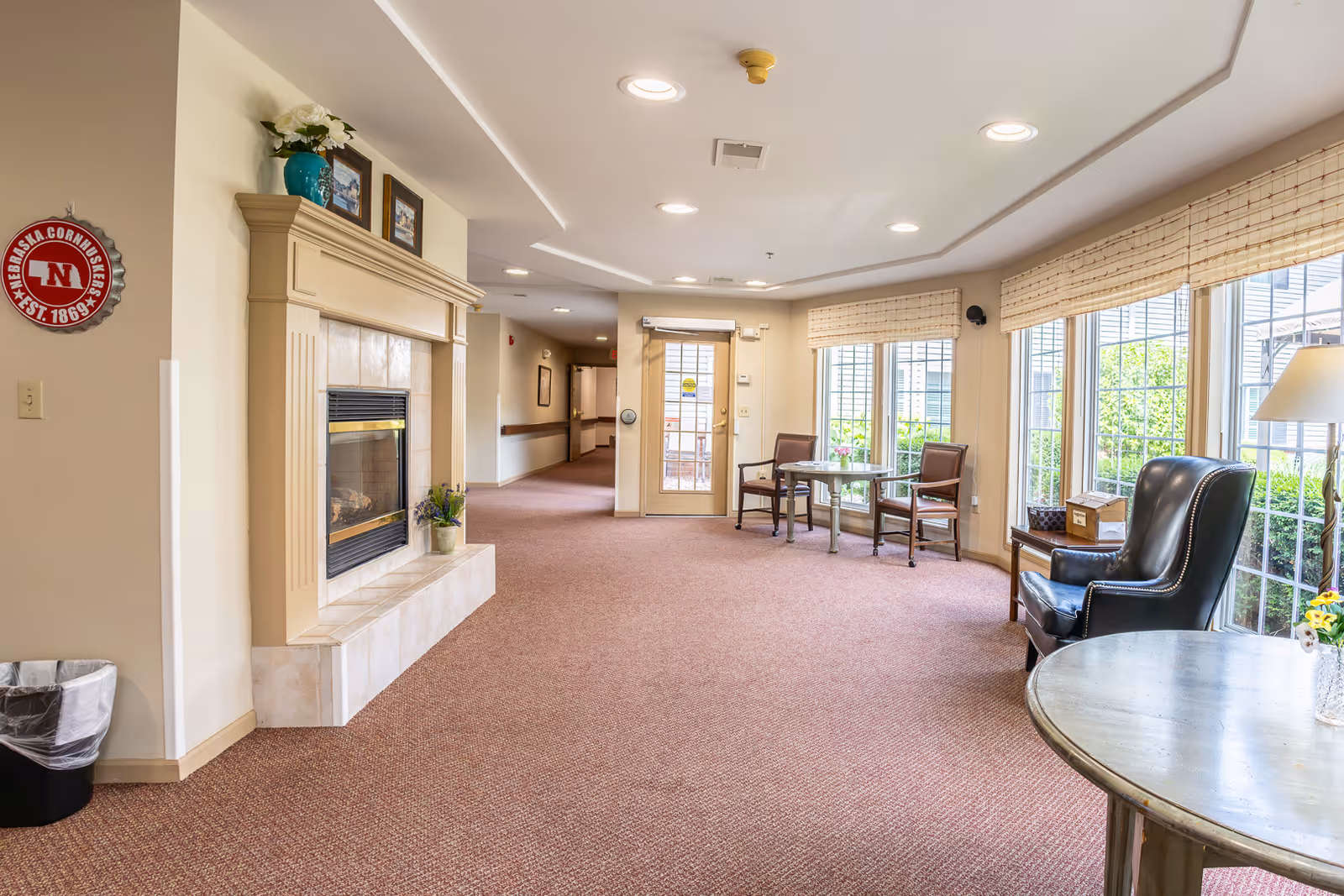 A bright and spacious common area in an assisted living facility featuring a fireplace with decorative items on the mantel, a seating area with two chairs and a small table near large windows, and a hallway leading to other rooms. The room has carpeted floors and beige walls with ceiling lights.