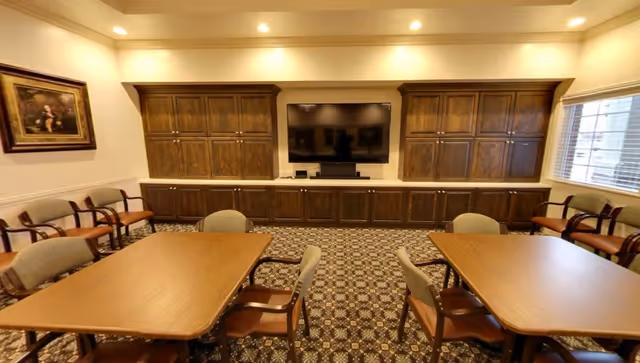 Communal dining/activity room with tables and chairs facing a wall of wooden cabinets and a mounted TV.