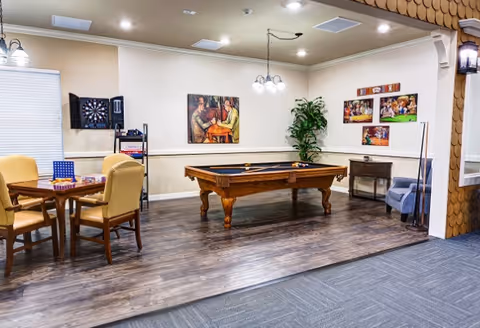 A recreational room featuring a pool table in the center, a small table with four chairs and board games on it, a dartboard on the wall, framed artwork, a potted plant, and a small cabinet with a blue armchair beside it.