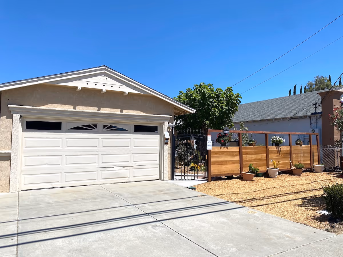 Exterior view of a residential building with a closed white garage door, a wooden fence with potted plants, and a gated entrance under a clear blue sky.