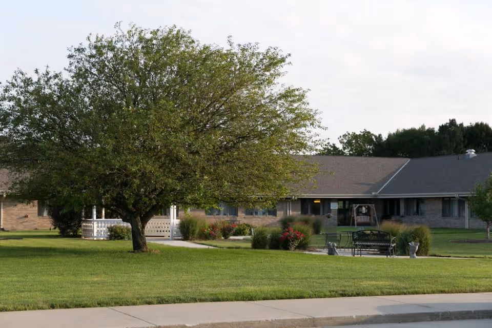 Single-story brick care facility building set behind a large tree with a manicured lawn, benches, and landscaping.