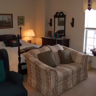 A cozy bedroom with a bed, a patterned loveseat with two cushions, a green armchair, a wooden dresser with a mirror, and a window letting in natural light. The room is softly lit by a table lamp on a nightstand beside the bed.