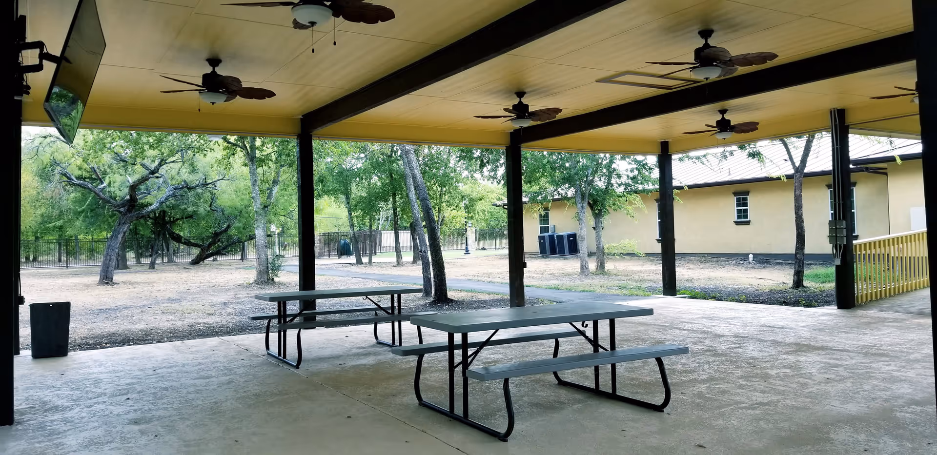 Covered outdoor patio area with two picnic tables and ceiling fans, overlooking a yard with trees and a building in the background.