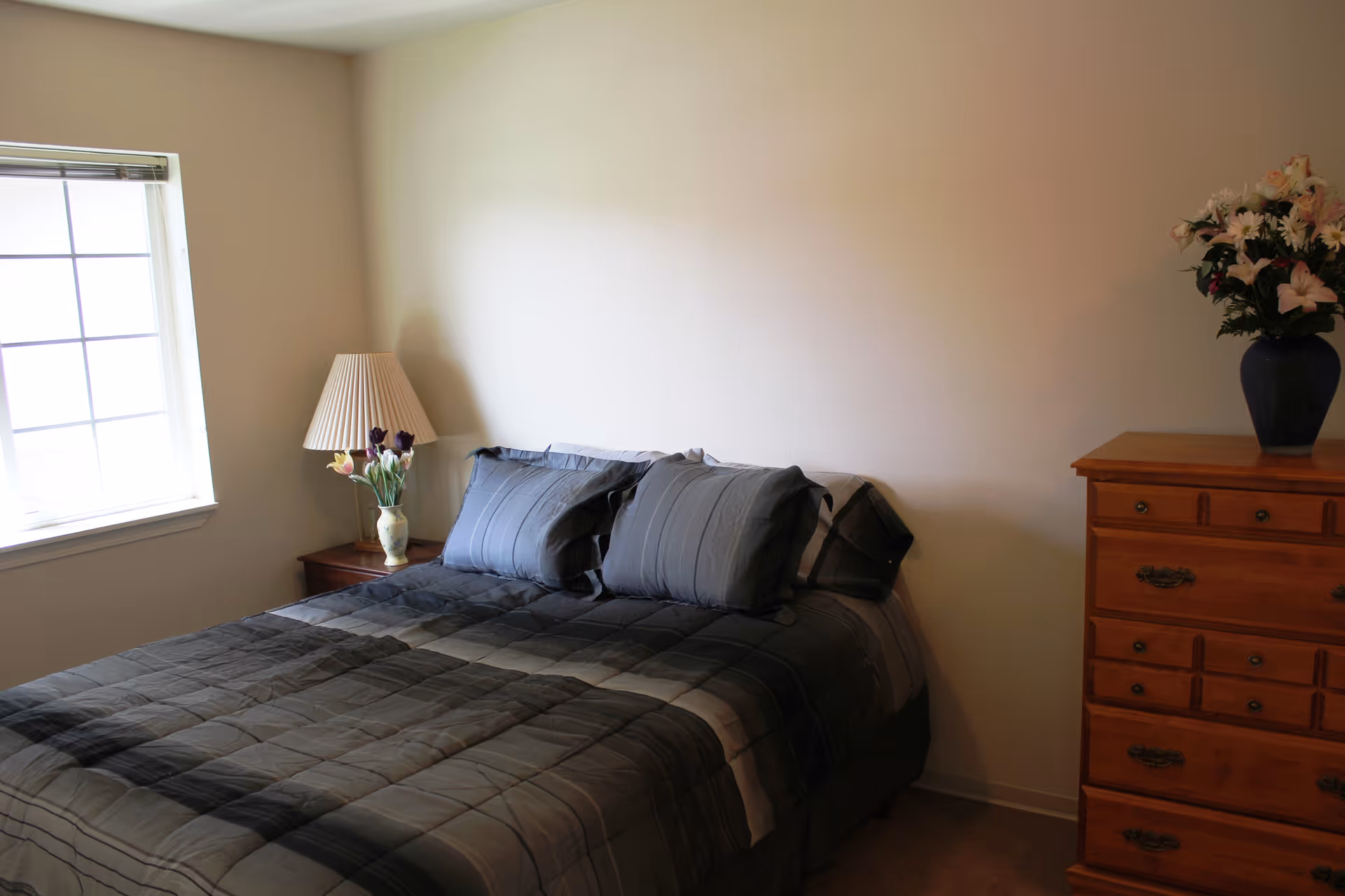A bedroom with a neatly made bed featuring a gray and black checkered comforter and matching pillows. To the left of the bed is a wooden nightstand with a lamp and a vase of flowers. To the right is a wooden dresser with a blue vase holding a bouquet of flowers. A window with blinds is on the left wall, allowing natural light into the room.