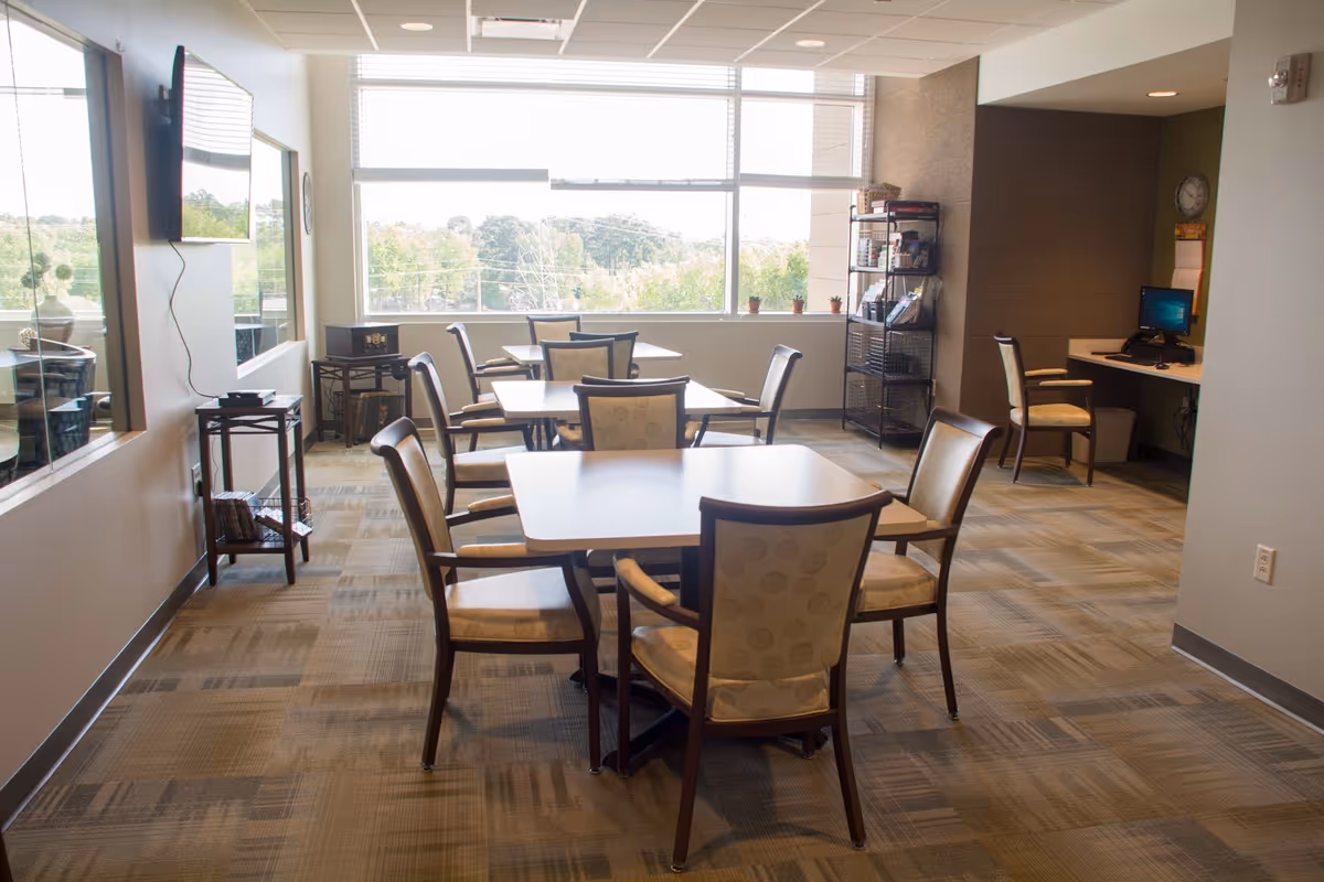 Bright communal dining/activity room with several tables and chairs and a computer workstation by a large window.