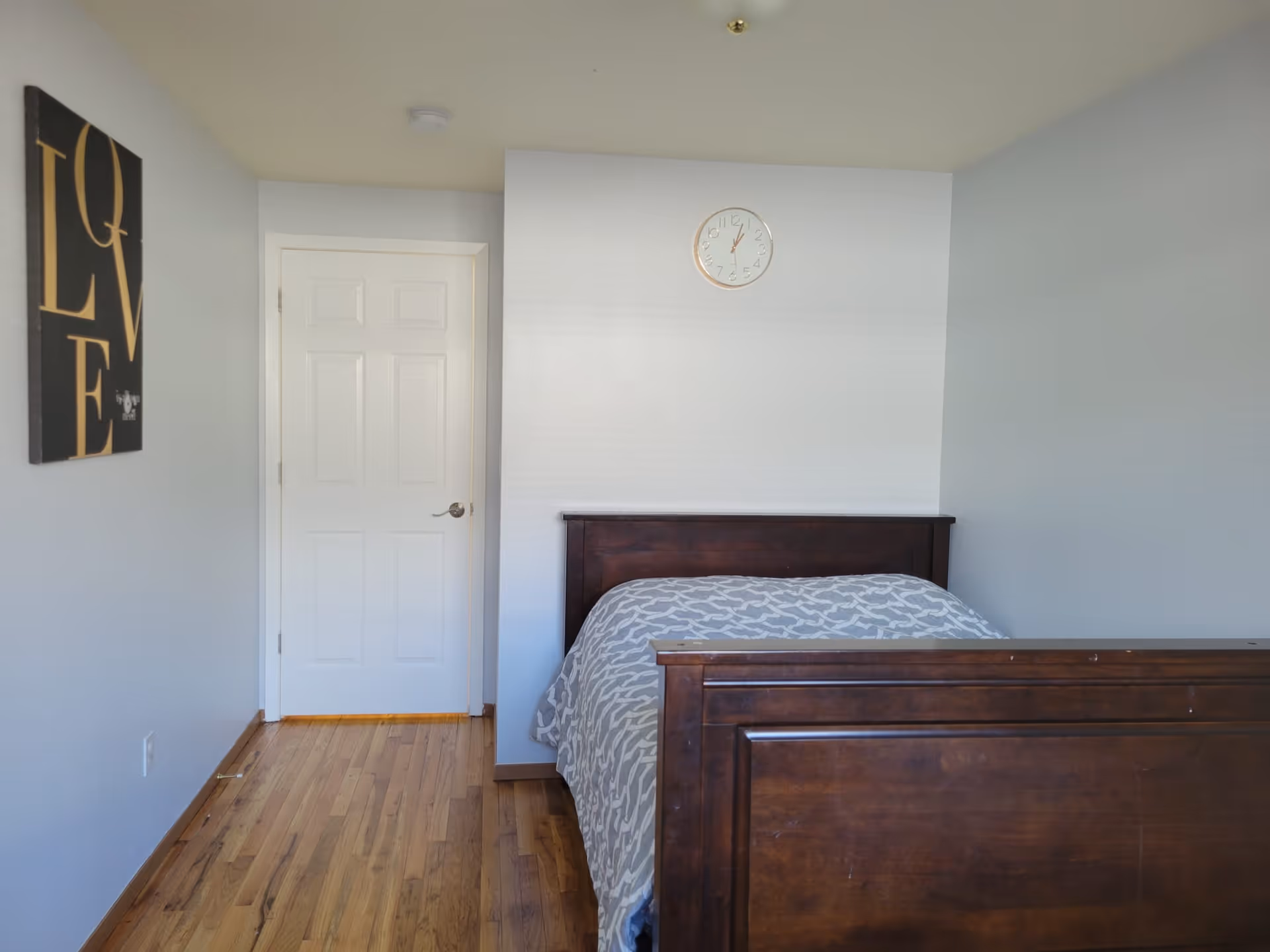A simple bedroom with a wooden bed frame and a gray patterned bedspread. There is a white door on the left wall, a clock above the bed on the back wall, and a wall decoration with the word 'LOVE' in large letters on the left wall. The floor is wooden.