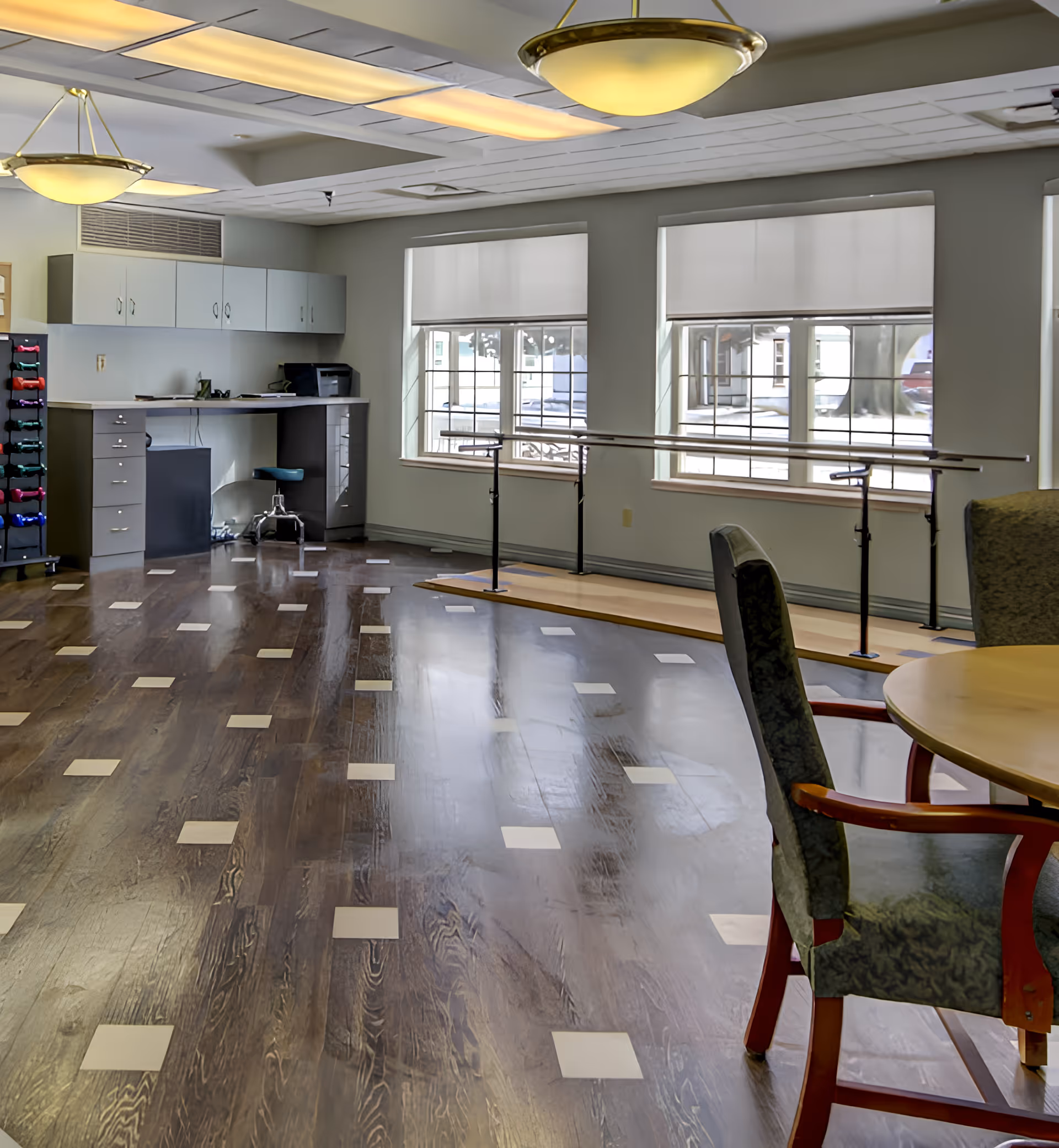 Interior room with wooden flooring featuring light square patterns, two large windows with white blinds, a ballet barre along the wall, a desk with cabinets and a chair, a rack of colorful dumbbells, and a round table with upholstered chairs.