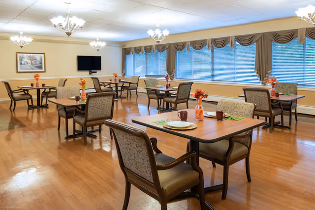 A spacious dining room with wooden floors and multiple tables set with plates, cups, and small flower vases. The room has large windows with blinds and brown valances, chandeliers hanging from the ceiling, and a wall-mounted television.