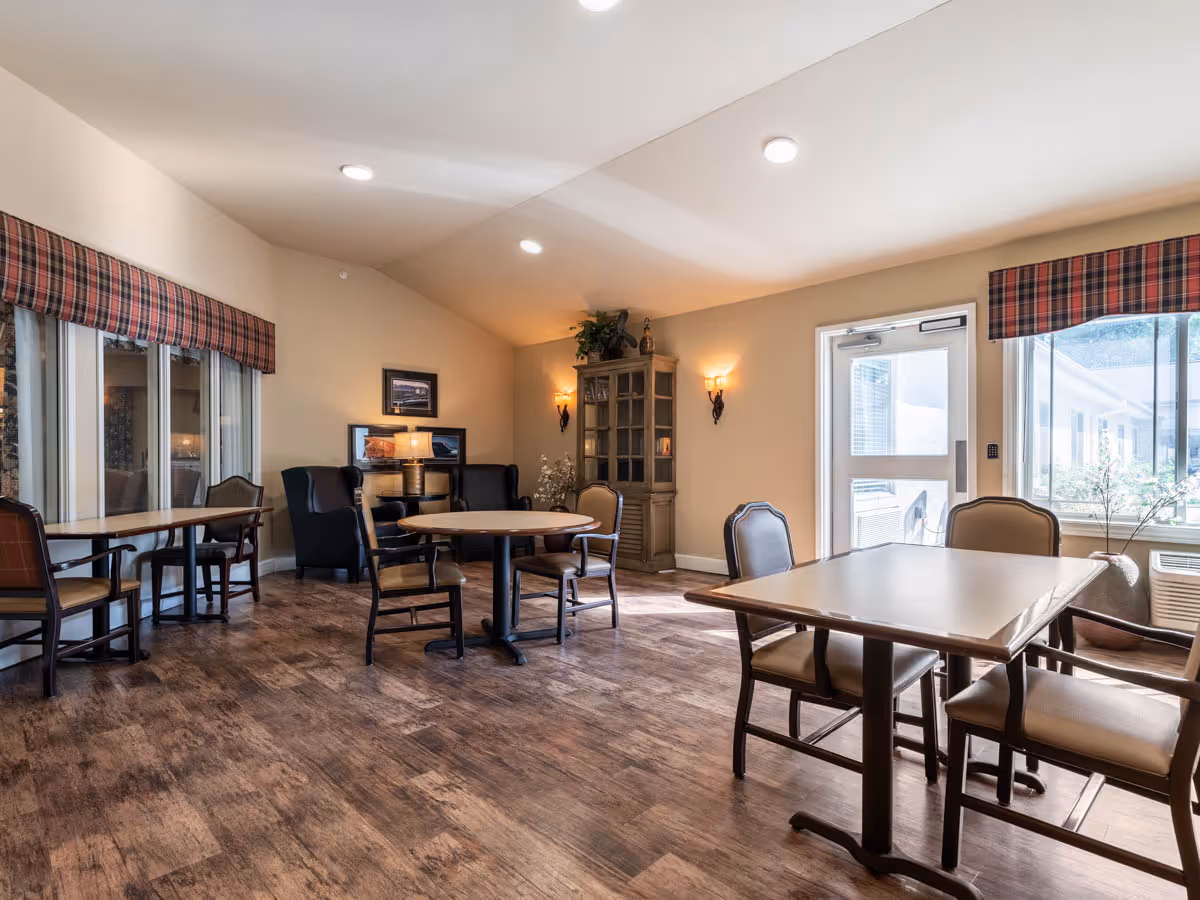 A well-lit common area with multiple tables and chairs arranged for seating. The room has wooden flooring, beige walls, and large windows with plaid valances allowing natural light to enter. There is a cabinet with decorative items and wall sconces providing additional lighting.