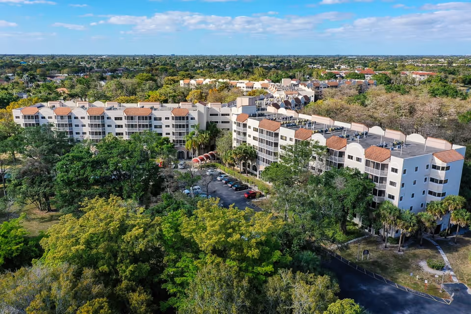 Aerial view of Forest Trace Senior Living facility showing multiple connected buildings with white walls and red-tiled roofs surrounded by trees and greenery. There is a parking lot with several cars visible in front of the buildings, and a suburban neighborhood can be seen in the background under a clear blue sky.