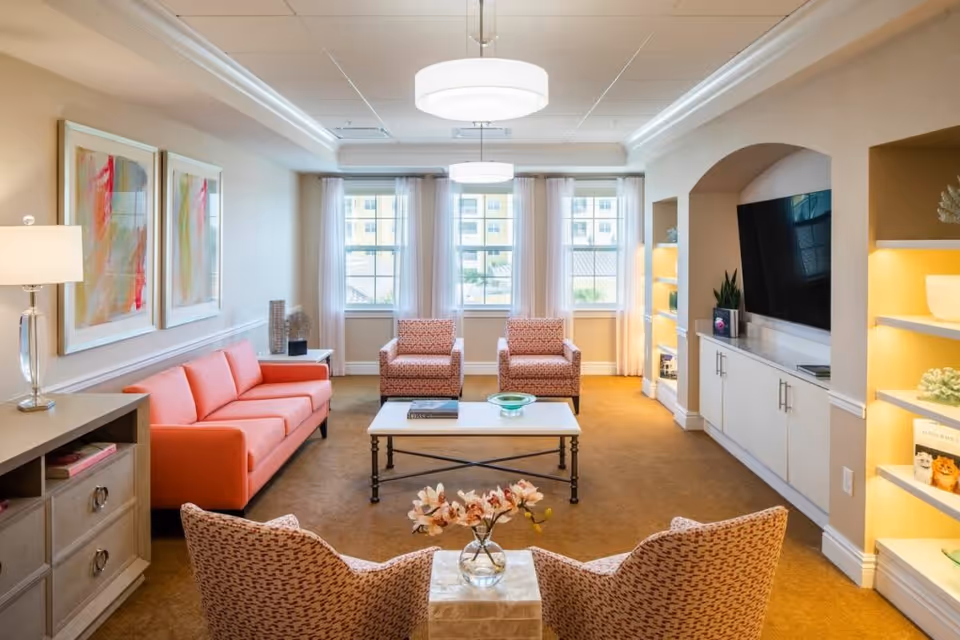A bright and cozy living room with a coral-colored sofa, two patterned armchairs facing a coffee table, and two more armchairs near the foreground. The room features large windows with sheer curtains, built-in shelves with decorative items, a flat-screen TV mounted in an alcove, and two modern ceiling lights. The walls are light beige, and the carpet is a warm brown color.
