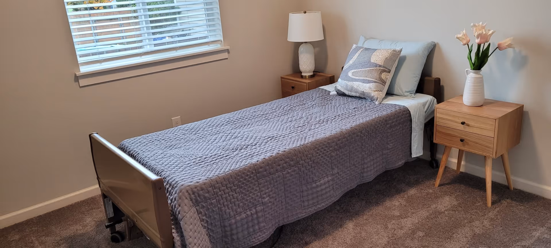 Small bedroom with a single bed covered in a gray quilt, two wooden nightstands, a lamp and a vase of flowers, and a window with blinds.