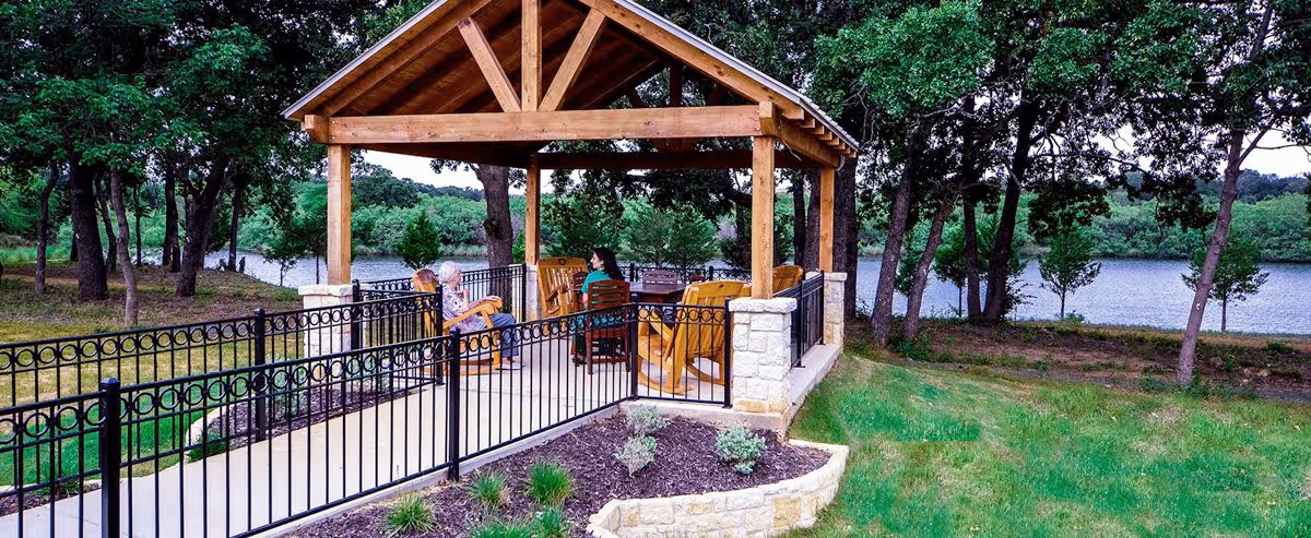 Outdoor covered seating area with wooden beams and stone pillars, featuring several people sitting on wooden chairs and benches. The area is surrounded by a black metal fence and overlooks a scenic view of trees and a body of water in the background.