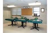 A room with three tables covered in green tablecloths, each with a small plant centerpiece. The room has a bulletin board on the wall, two windows with blinds, and a clock above the bulletin board. There are cabinets along one wall and a framed picture on another wall.