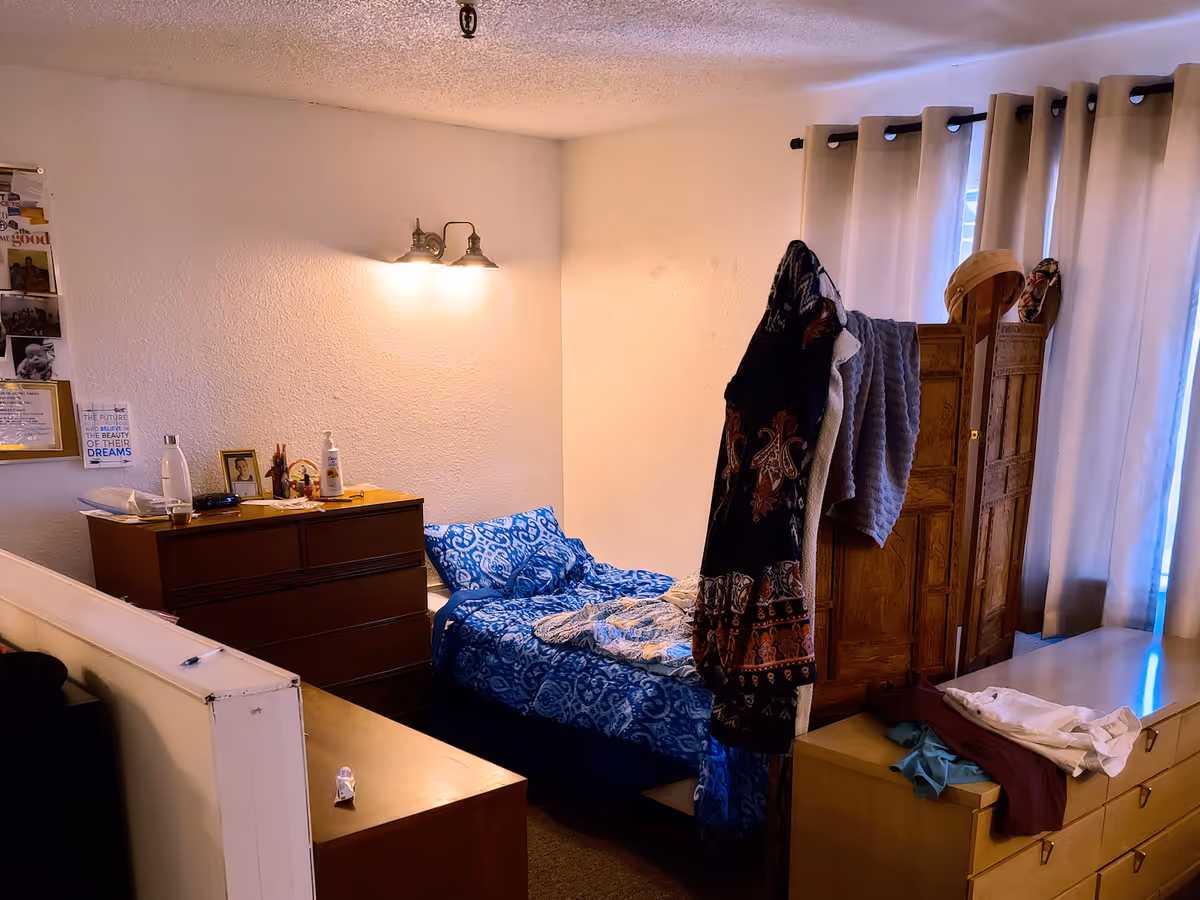 A small bedroom with a single bed covered in blue patterned bedding. There is a wooden dresser with various items on top, including a water bottle, lotion, and framed photos. A wall-mounted light fixture illuminates the room. Clothes and towels hang on a wooden folding screen near a window with beige curtains. Another dresser with folded clothes is visible in the foreground.