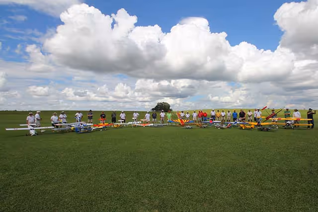 A large group of people standing in a line behind colorful model airplanes on a grassy field under a partly cloudy sky.