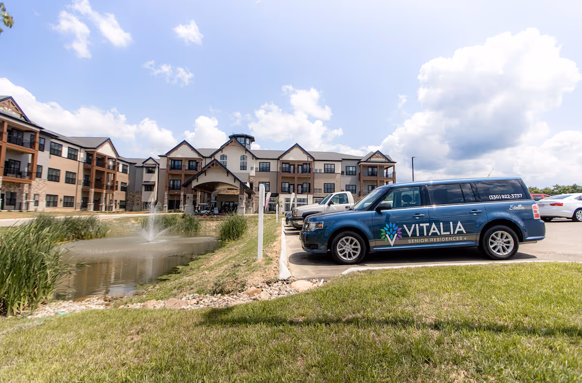 Exterior view of Vitalia Senior Residences at Stow, showing a large multi-story building with balconies, a pond with a fountain, and a parking lot with a blue vehicle branded with the Vitalia Senior Residences logo.
