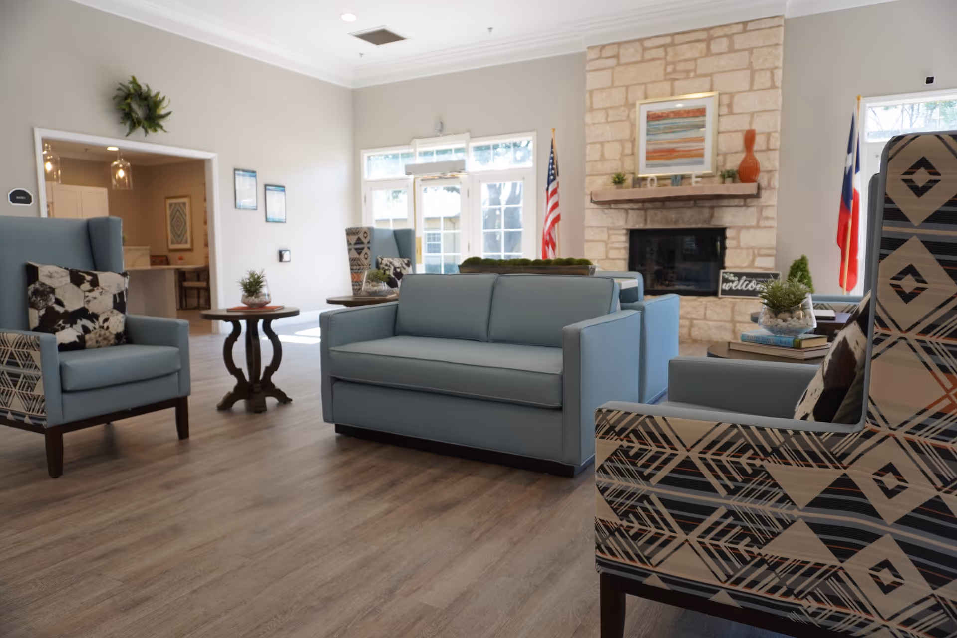 Sunny common living area with blue sofas and patterned armchairs arranged around tables facing a stone fireplace.
