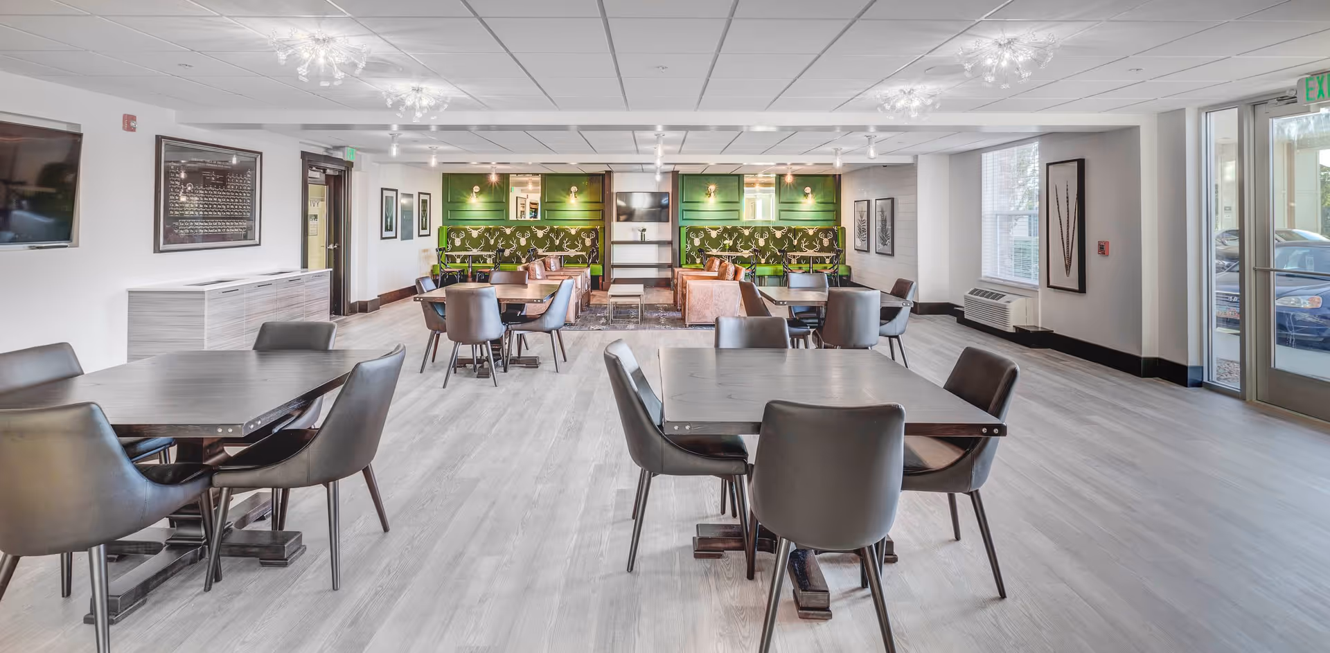 A bright and modern common area with multiple dark wood tables and black chairs arranged for seating. The back wall features green paneling with patterned upholstery and leather armchairs, along with a mounted TV. Large windows and glass doors allow natural light to fill the space, which has light wood flooring and contemporary ceiling lights.