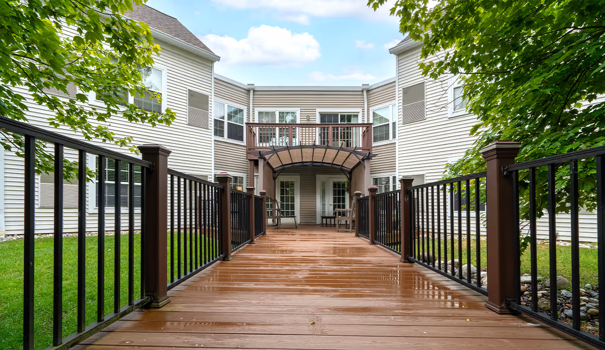 View of a wooden deck with black metal railings leading to the entrance of a two-story building with beige siding. The building has multiple windows and a small balcony above the entrance. Green trees and grass surround the deck on both sides under a partly cloudy sky.
