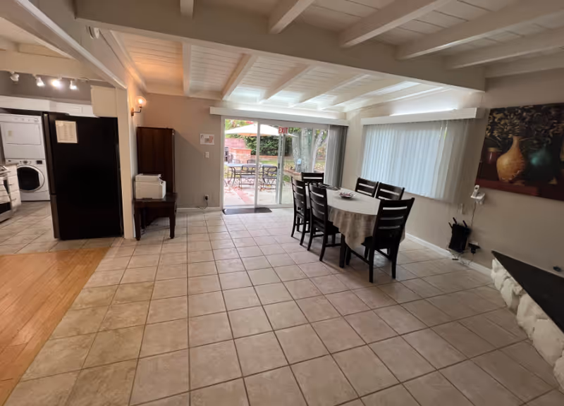 Interior view of a dining area in an assisted living facility with a rectangular table covered by a tablecloth and six chairs around it. The room has tiled flooring, a large window with vertical blinds, and a sliding glass door leading to an outdoor patio with tables and umbrellas. To the left, there is a kitchen area with a refrigerator and a stacked washer and dryer. The ceiling has exposed beams and the walls are painted light gray. A painting of vases hangs on the right wall.
