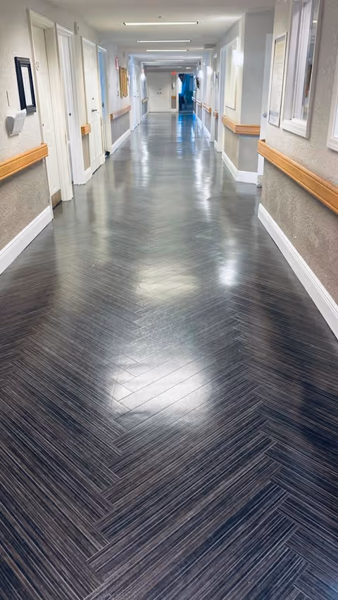A long, clean hallway inside a skilled nursing facility with dark herringbone patterned flooring, beige walls, wooden handrails on both sides, and several white doors along the corridor. The hallway is well-lit with ceiling lights and has framed pictures on the walls.