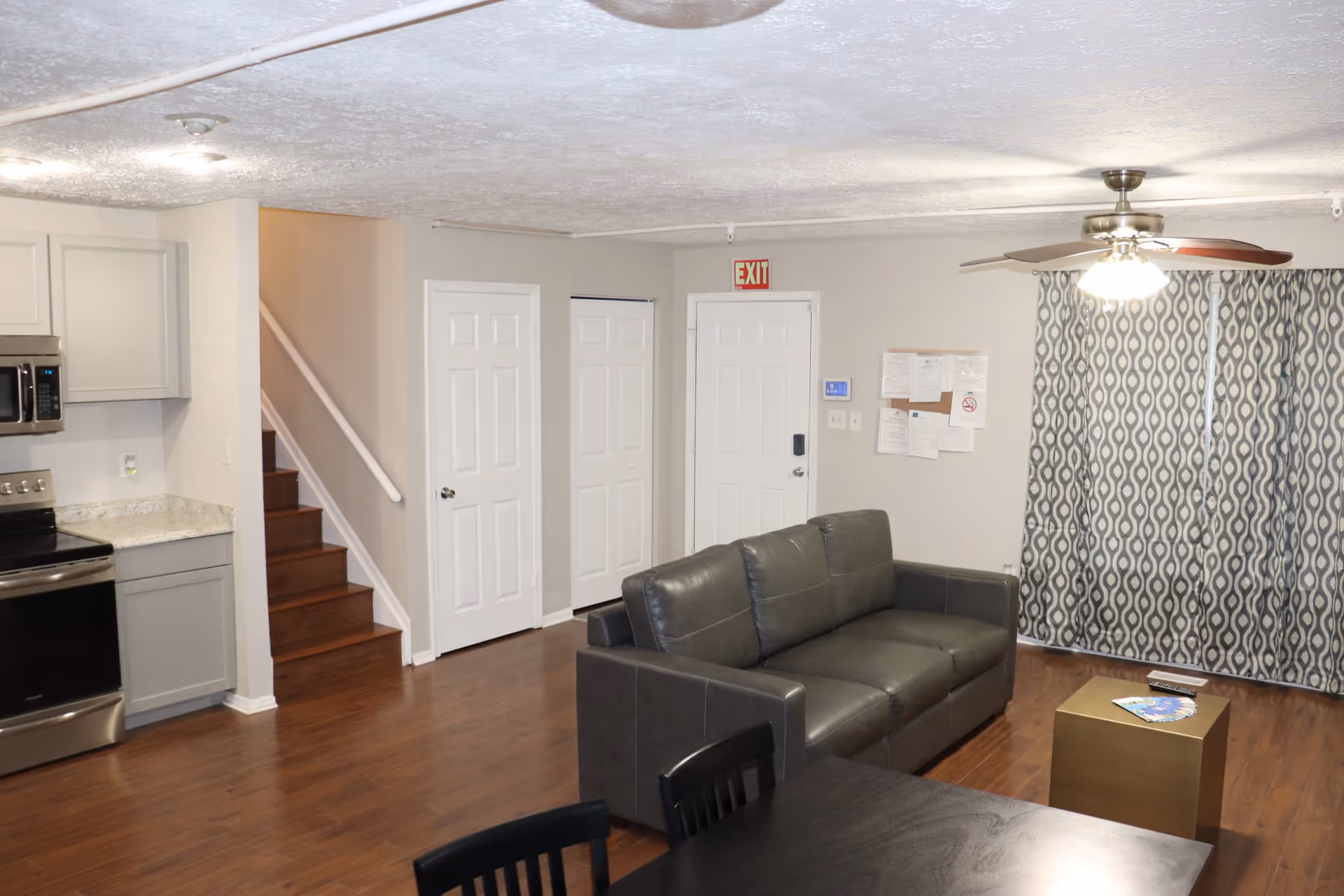 Interior view of a living area with a gray leather sofa, a small gold coffee table with magazines and a remote control, a ceiling fan with lights, patterned curtains, and a kitchen area with stainless steel appliances and gray cabinets. There are wooden stairs and multiple white doors along the wall.