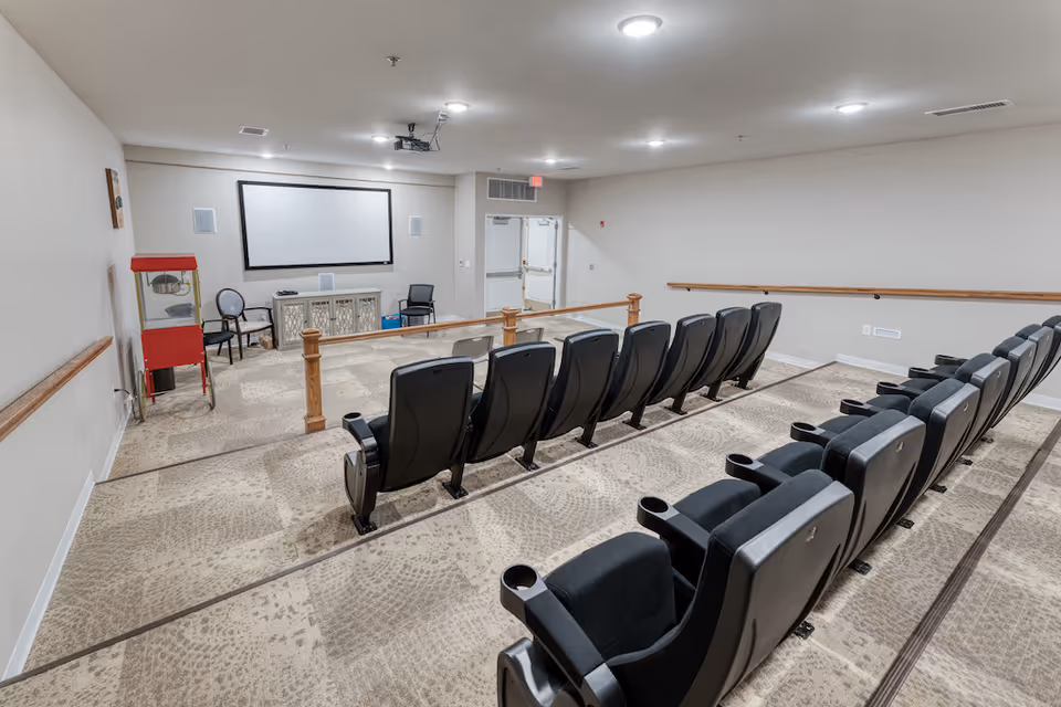 A small theater room with two rows of black reclining seats facing a large white projection screen mounted on the wall. There is a red popcorn machine on the left side near the wall, two chairs, and a cabinet below the screen. The room has beige carpet with a patterned design, white walls, and ceiling lights.