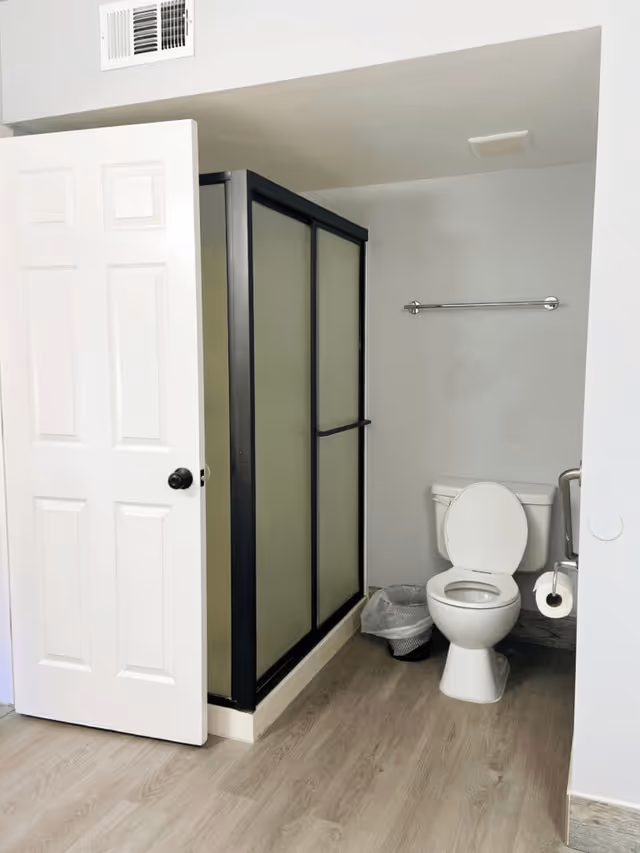 A bathroom with a white toilet, a trash can beside it, a towel rack on the wall, and a shower with frosted glass sliding doors and a black frame. The door to the bathroom is open, and the floor has light wood-style flooring.